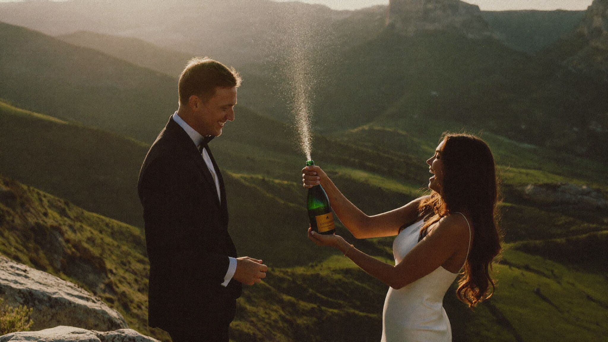 Couple popping champagne on a cliff edge at sunset after their elopement ceremony