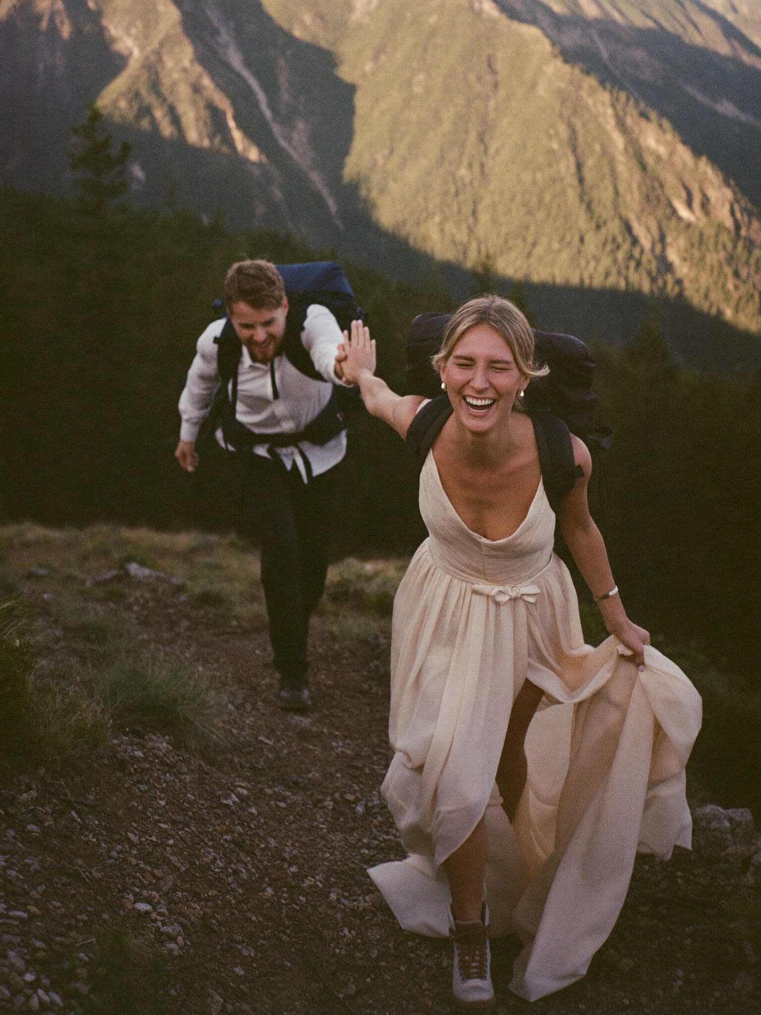 Bride in wedding dress and hiking boots on a mountain trail with her groom, adventure elopement