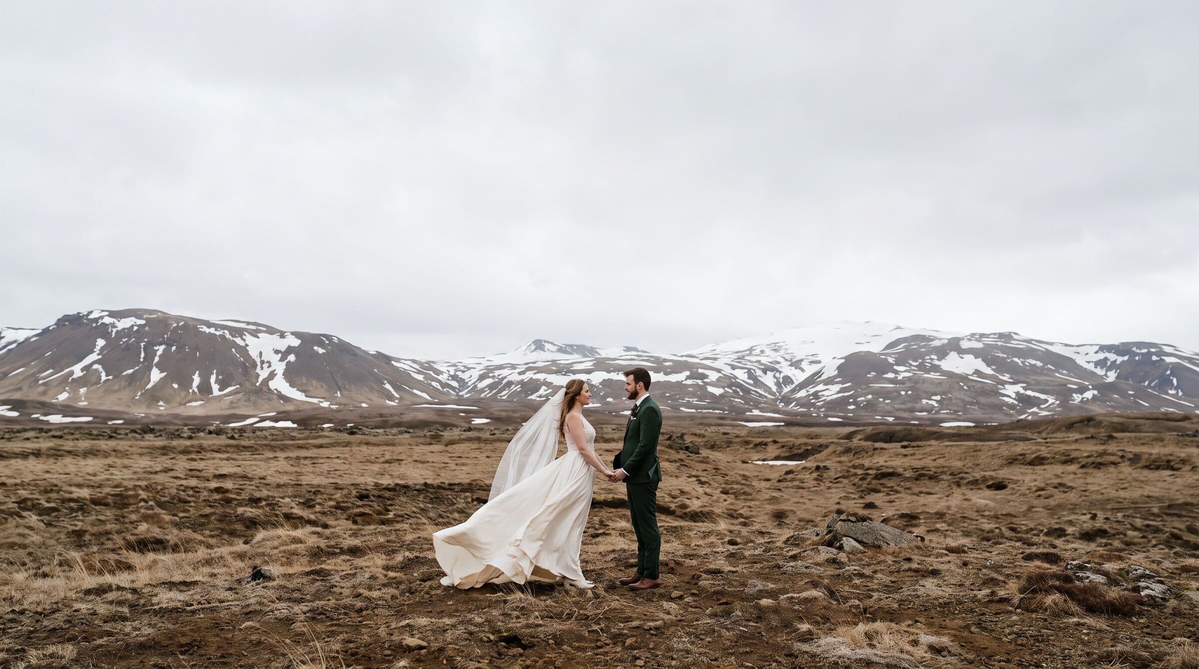 Bride and groom holding hands in an Icelandic lava field with snow-capped mountains