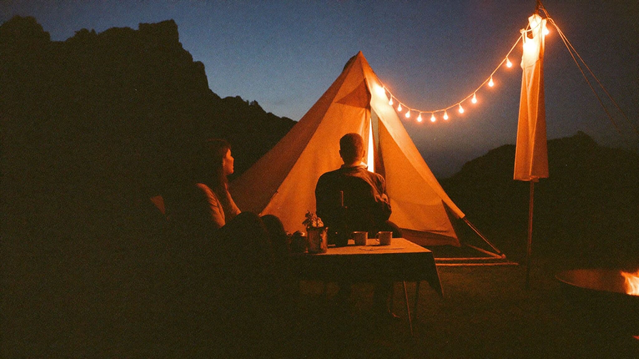 Couple relaxing at their tent camp at dusk after their mountain elopement, lanterns glowing against the ridgeline